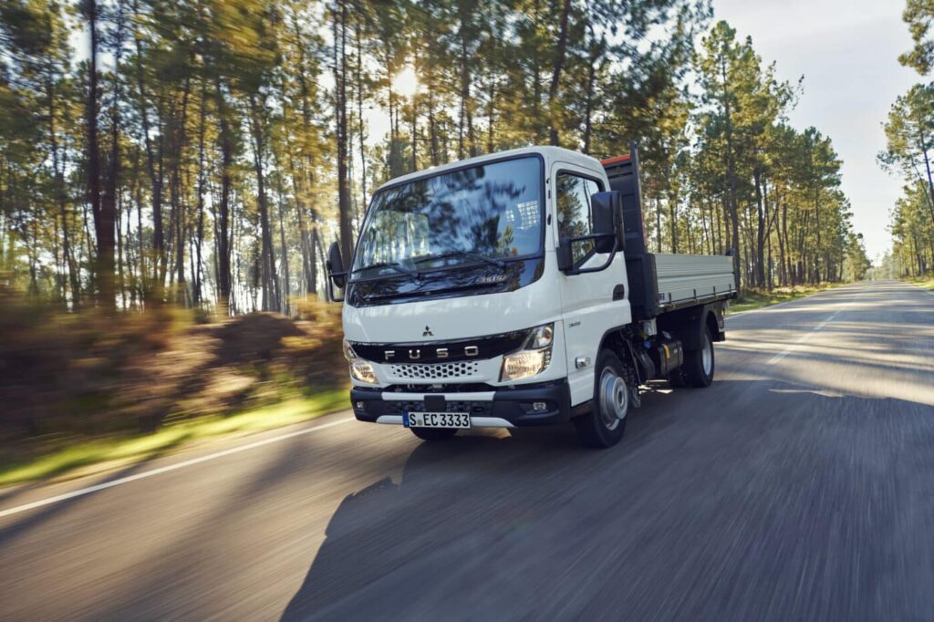 Ein weißer FUSO Canter 3,5-Tonner fährt mit mittlerer Geschwindigkeit auf einer Landstraße, die durch einen sonnendurchfluteten Wald führt.