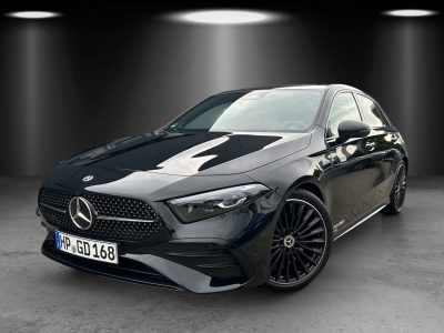 Front- und Seitenansicht eines schwarzen Mercedes-Benz Kompaktwagens in einem Studioumfeld mit Spotlights. Fahrzeug mit Diamantgrill, LED-Scheinwerfern, AMG-Line Frontschürze und schwarzen mehrspeichigen Leichtmetallrädern.
