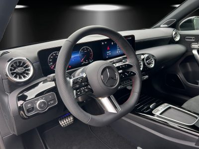Innenraum eines modernen Mercedes-Benz mit AMG-Line Ausstattung. Sicht auf das perforierte Sportlenkrad mit roten Kontrastnähten, volldigitales Kombiinstrument mit zwei Displays, runde Lüftungsdüsen im Turbinen-Design sowie Mittelkonsole in Klavierlackoptik. Ambiente-Beleuchtung in Blau dezent im Hintergrund.