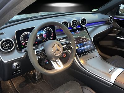 Cockpit eines modernen Mercedes-AMG mit digitalem Kombiinstrument, großem Touchscreen in der Mittelkonsole und Ambientebeleuchtung in Blau. Sicht auf sportliches Multifunktionslenkrad mit AMG-Logo, Aluminium-Details und Carbon-Dekor.