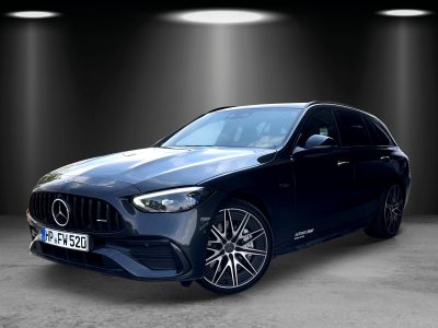 Dunkler Mercedes-Benz Kombi mit sportlicher AMG-Frontschürze und markanten Leichtmetallfelgen, schräg von vorne fotografiert. Das Fahrzeug steht in einer ausgeleuchteten Studio-Umgebung.