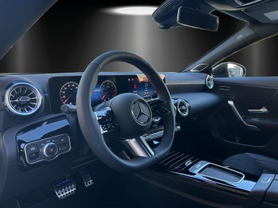 Mercedes-Benz Interieuransicht aus Fahrersicht. Sportlenkrad mit roter Kontrastnaht, volldigitales Cockpit mit Navigationsanzeige, runde Lüftungsdüsen im Turbinenlook, Ambientebeleuchtung in Blau, Aluminium-Sportpedale, schwarze Hochglanz-Mittelkonsole, Sitze in Mikrofaser und Lederoptik mit roten Nähten.