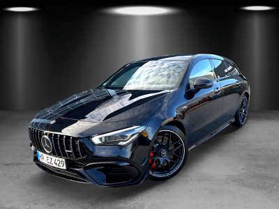 Front- und Seitenansicht eines schwarzen Mercedes-AMG CLA 45 S Shooting Brake mit Panamericana-Kühlergrill, LED-Scheinwerfern, roten Bremssätteln und schwarzen AMG-Felgen. Das Fahrzeug steht in einem Studio mit Spotbeleuchtung auf grauem Betonboden.