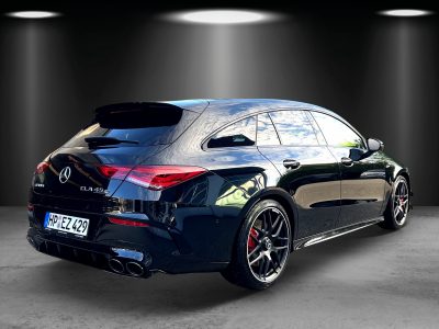 Rückansicht eines schwarzen Mercedes-AMG CLA 45 S Shooting Brake mit vierflutiger Abgasanlage, getönten Scheiben, schwarzen Leichtmetallfelgen und roten Bremssätteln. Das Fahrzeug steht in einem Studio-Setup mit Spotbeleuchtung auf grauem Boden.