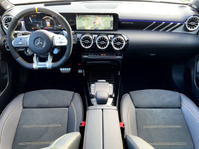 Cockpit-Ansicht eines Mercedes-AMG mit digitalem Kombiinstrument, Navigationsbildschirm, beleuchteter Zierleiste und drei runden Lüftungsdüsen in der Mittelkonsole. AMG Performance-Lenkrad mit gelbem Mittenmarker und Sportsitze mit gelben Ziernähten sichtbar.
