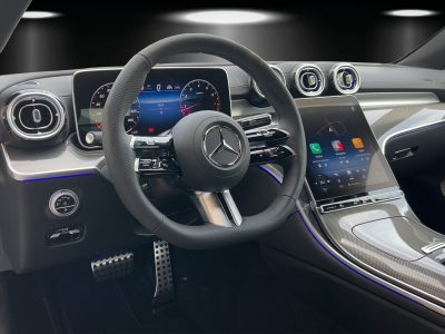 Detailaufnahme des Innenraums eines Mercedes-Benz – AMG-Multifunktionslenkrad mit perforiertem Leder, digitale Cockpit-Anzeige, großes zentrales Touchdisplay mit Navigationssystem, Ambientebeleuchtung in Blau, Aluminium- und Carbon-Elemente, sportliche Pedalerie.