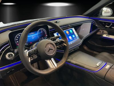 Innenraum eines Mercedes-Benz mit digitalem Cockpit, großem zentralen Touchscreen, AMG-Lenkrad, Ambientebeleuchtung in Blau und modernen Designelementen – Fokus auf Fahrerperspektive.