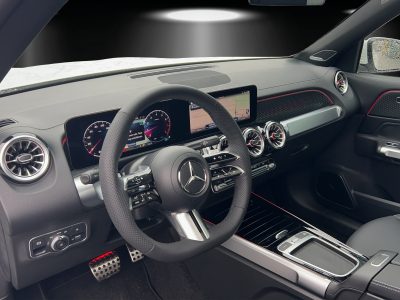 Innenraum eines Mercedes-Benz mit sportlichem Multifunktionslenkrad, digitalem Cockpit, zentralem Navigationsdisplay, Aluminium-Applikationen und roten Ziernähten.