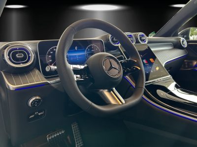 Innenraum eines Mercedes-Benz mit modernem Multifunktionslenkrad, volldigitalem Cockpit und großem zentralem Touchscreen – Ambientebeleuchtung in Blau unterstreicht das elegante Design.