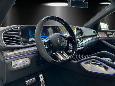 Innenraum eines modernen Fahrzeugs in Studiobeleuchtung, Blick vom Fahrersitz auf ein Multifunktionslenkrad mit Alcantara-Bezug, digitales Cockpit, Ambientebeleuchtung in Blau sowie Karbonelemente am Armaturenbrett und an der Tür.